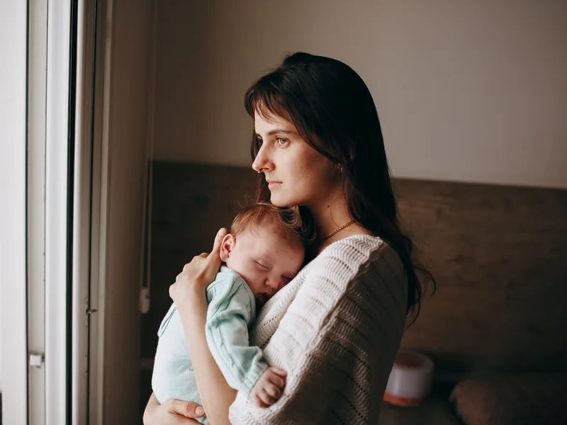 A mother standing by a window, holding her sleeping newborn against her shoulder